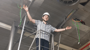 Photo of a person in an industrial setting high up on gantry wearing a white hard hat with their arms out stretched
